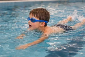 Kisgyermek oldalnézetből, amint önállóan mellúszásban úszik egy beltéri uszodában, úszószemüvegben, természetes mozgás közben.