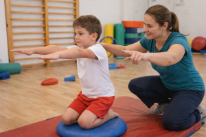 Gyógytestnevelő szakember játékos mozgásgyakorlat közben segít egy kisfiúnak egyensúlyozó párnán egy világos tornateremben, a törzs- és tartásjavítás fejlesztése érdekében.