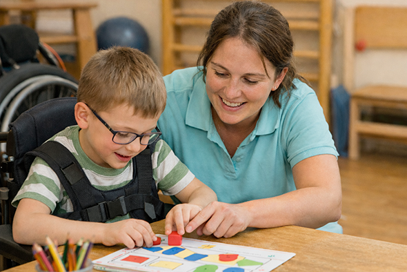 Konduktor szakember segít egy mozgásában korlátozott kisfiúnak az önálló járás gyakorlásában speciális korlátok között a Fővárosi Pedagógiai Szakszolgálat fejlesztő termében.