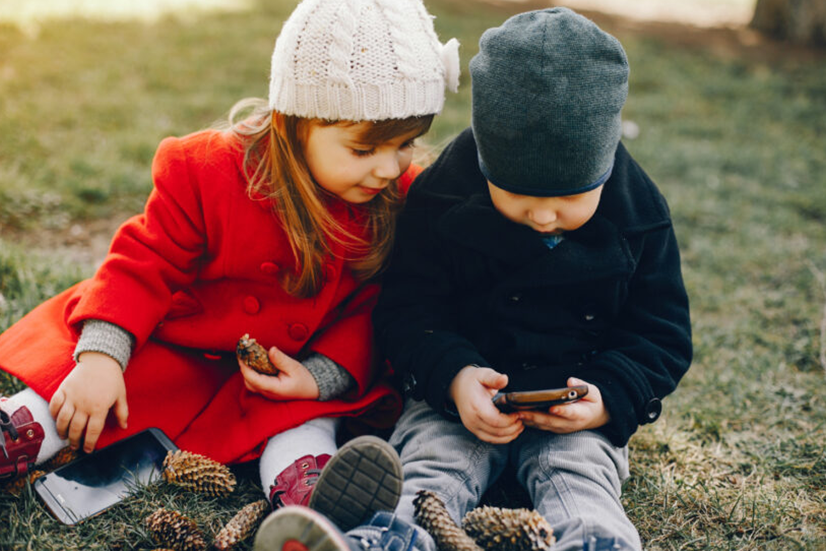 Egy kisgyermek a szabadban, egy parkban ülve néz egy mobileszközt, szemléltetve a digitális eszközök jelenlétét a mindennapi környezetben.
